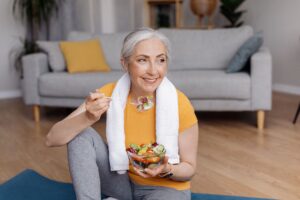 Woman in yellow t-shirt with towel around her neck eating a salad on her yoga mat